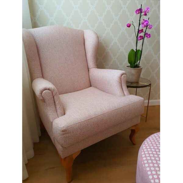 כורסה מעוצבת בצבע ורוד בהיר, לצד צמח פרחוני, על רקע טפט עם דגם גיאומטרי.